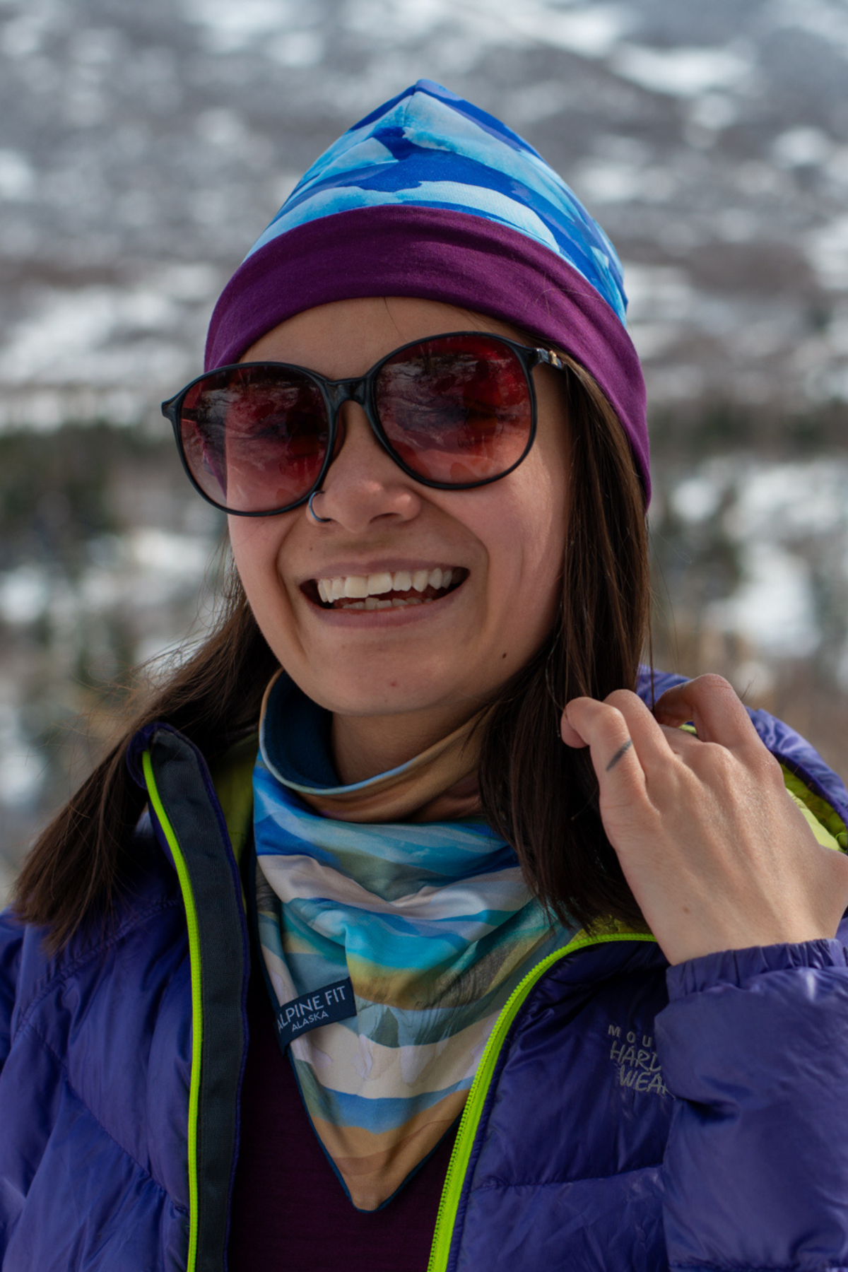 Alpine Fit Go All Winter Merino Wool Hat Glacial, Person wearing a colorful beanie, sunglasses, and a blue jacket with a snowy background
