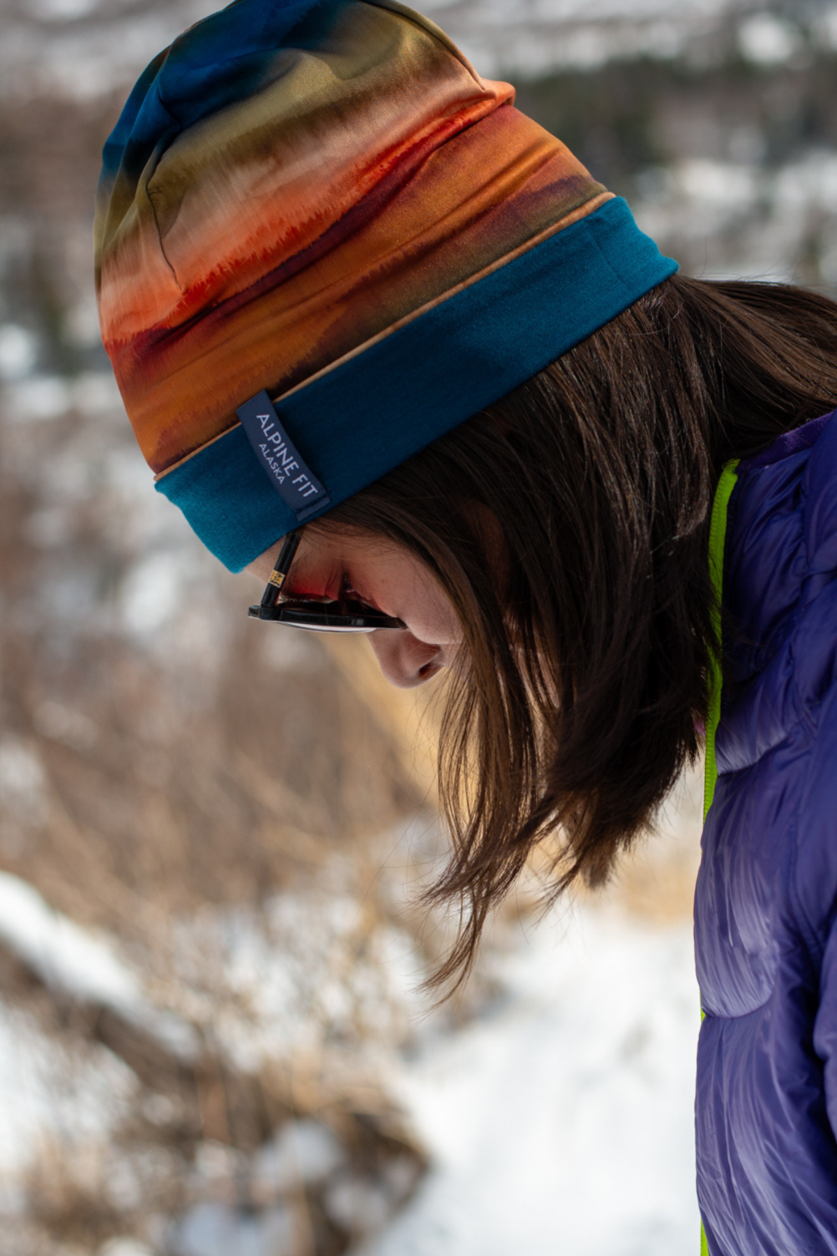 Alpine Fit Go All Winter Merino Wool Hat Grasslands, Person wearing a colorful beanie and sunglasses in a snowy landscape