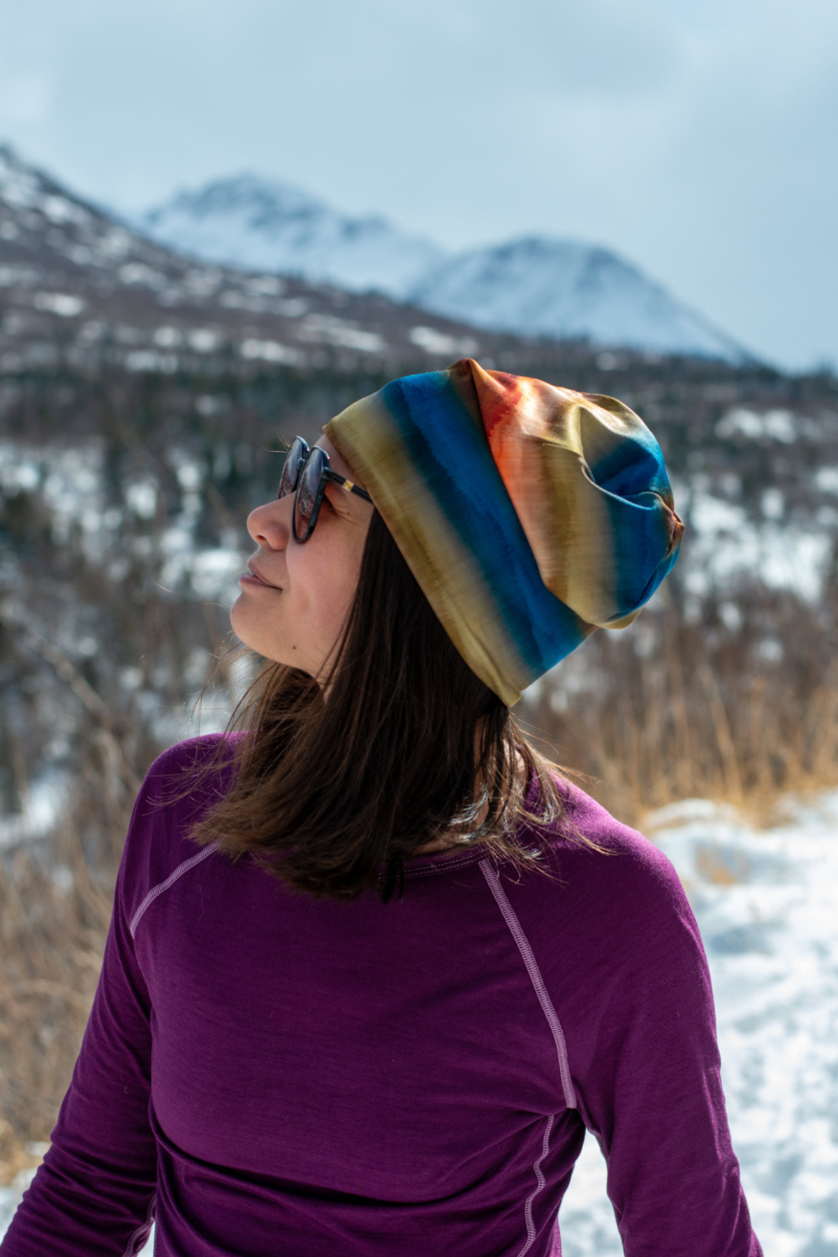 Alpine Fit Go All Winter Merino Wool Hat Grasslands, Person wearing a colorful striped beanie in a snowy mountain landscape