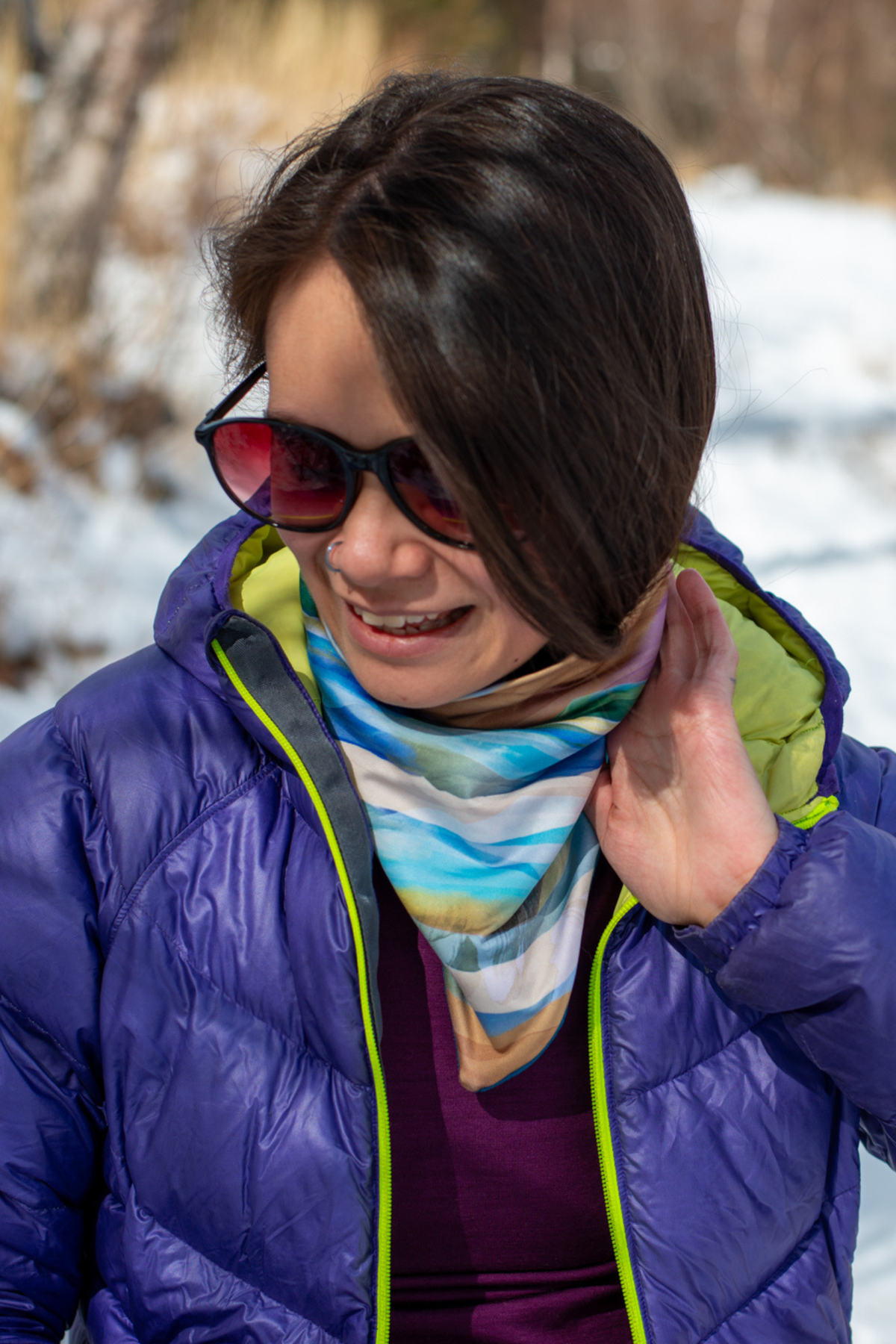 Alpine Fit Mountain V Fleece Neck Warmer Rainbow Peak, Woman wearing a purple jacket and colorful neck warmer outdoors with snow and trees in the background