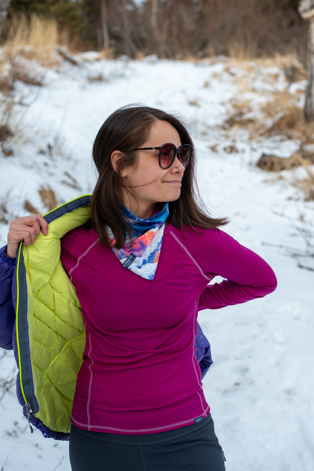 Alpine Fit Mountain V Merino Wool Neck Warmer Sunrise, Woman in a snowy landscape wearing a purple shirt, green vest, and sunglasses.