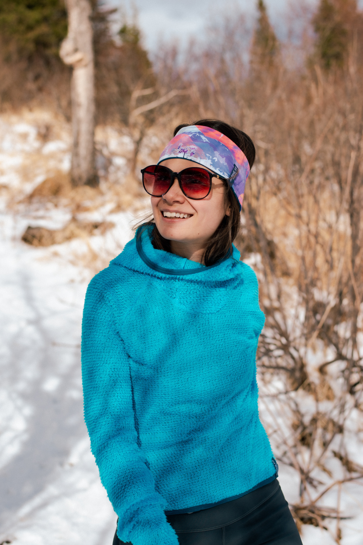 Alpine Fit Trailhead Cozy Fleece Headband Sunrise, Person wearing a blue sweater and colorful headband in a snowy outdoor setting