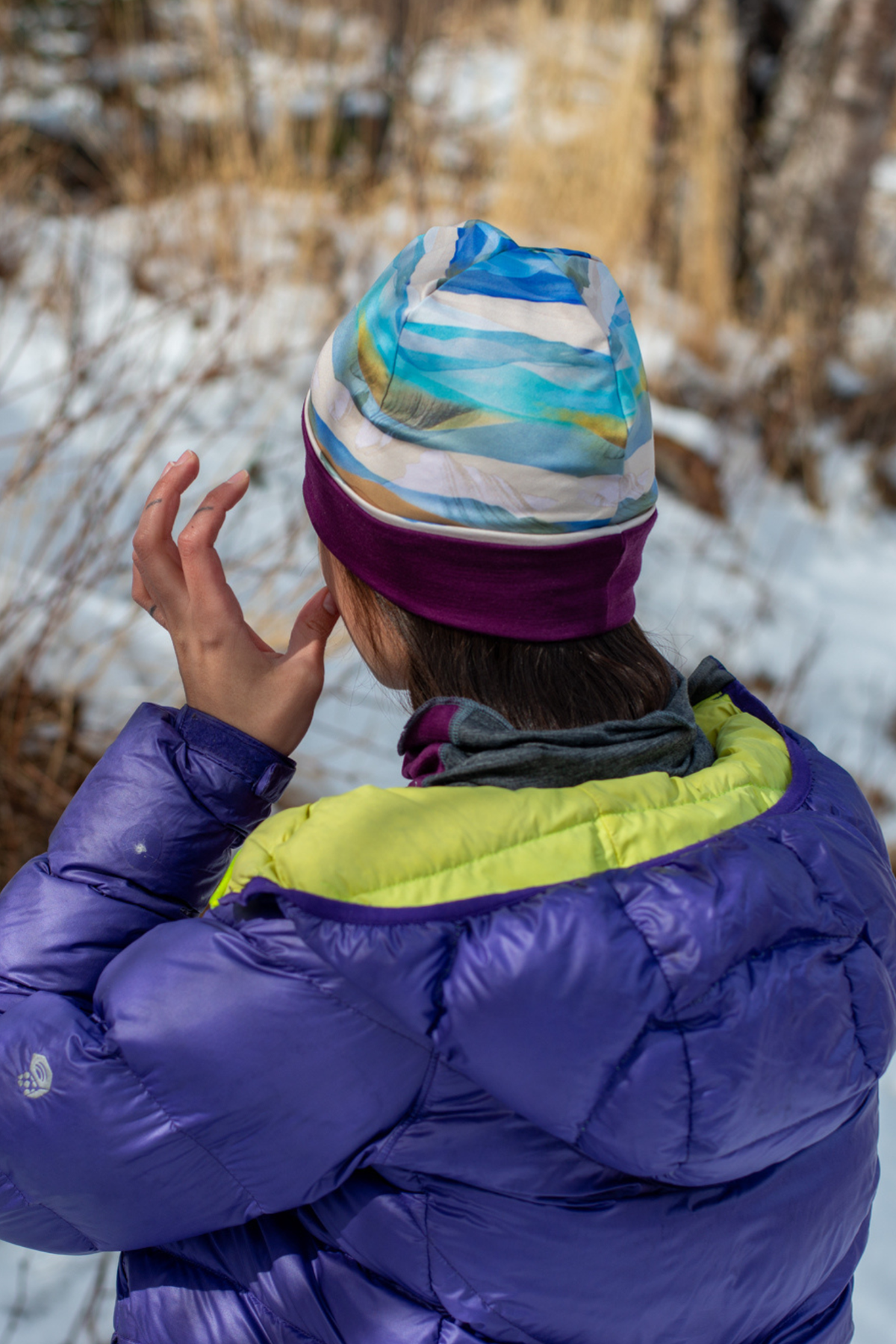 Alpine Fit Go All Winter Merino Wool Hat Rainbow Peak, Person wearing a colorful striped beanie and purple jacket in a snowy forest.