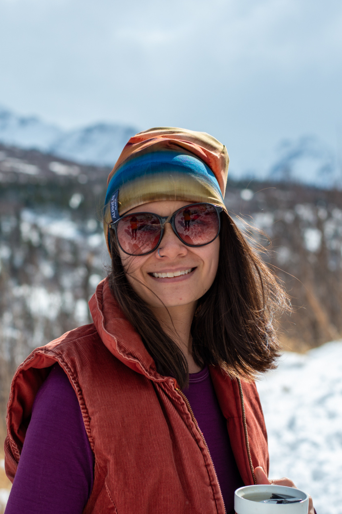 Alpine Fit Go All Winter Merino Wool Hat Grasslands, Person wearing sunglasses and a red vest in a snowy landscape with mountains.